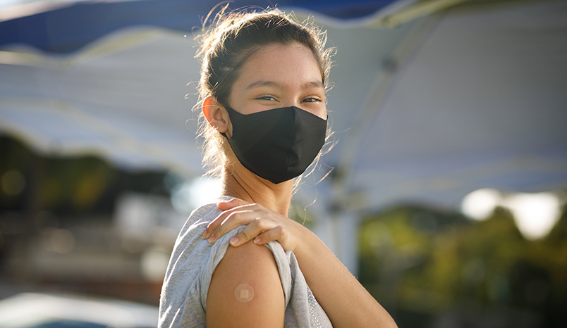 Woman with face mask lifts sleeve to show band-aid.