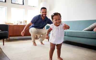 Baby walks in living room while dad watches.