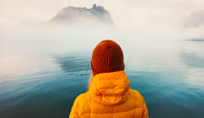 Woman in winter coat looks out onto foggy waters.